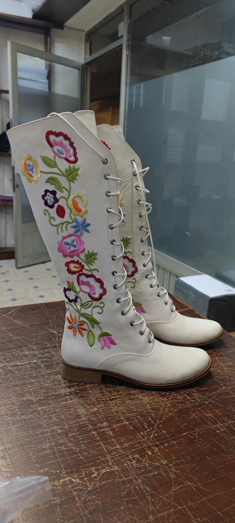 May include: White knee-high boots with colorful floral embroidery. The boots feature a lace-up design and a low heel. The floral design includes pink, blue, yellow, and orange flowers with green leaves. The boots are displayed on a wooden surface.