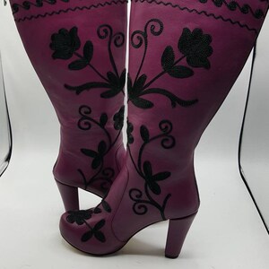 Puede incluir: Un par de botas altas de cuero morado con bordados florales negros y dise&ntilde;os de enredaderas. Las botas tienen punta redondeada, cierre de cremallera lateral, un tac&oacute;n de bloque resistente y una l&iacute;nea negra ondulada decorativa arriba.