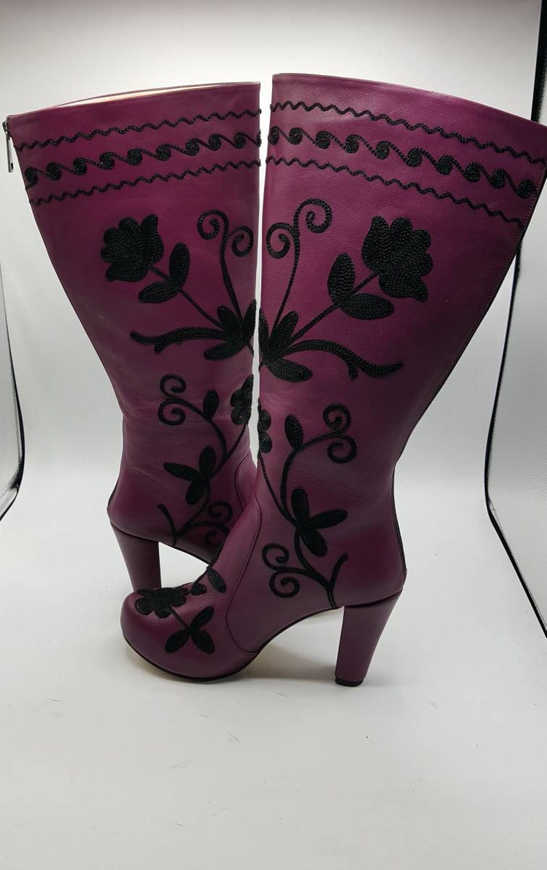 Puede incluir: Un par de botas altas de cuero morado con bordados florales negros y dise&ntilde;os de enredaderas. Las botas tienen punta redondeada, cierre de cremallera lateral, un tac&oacute;n de bloque resistente y una l&iacute;nea negra ondulada decorativa arriba.