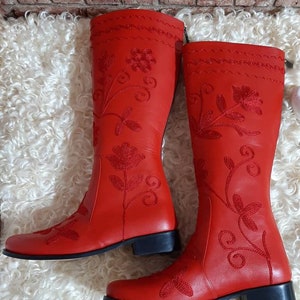 Puede incluir: Un par de botas rojas brillantes hasta la rodilla. Las botas presentan intrincados bordados florales en hilo rojo m&aacute;s oscuro. Tienen un tac&oacute;n bajo y negro y se muestran sobre una superficie blanca y esponjosa.