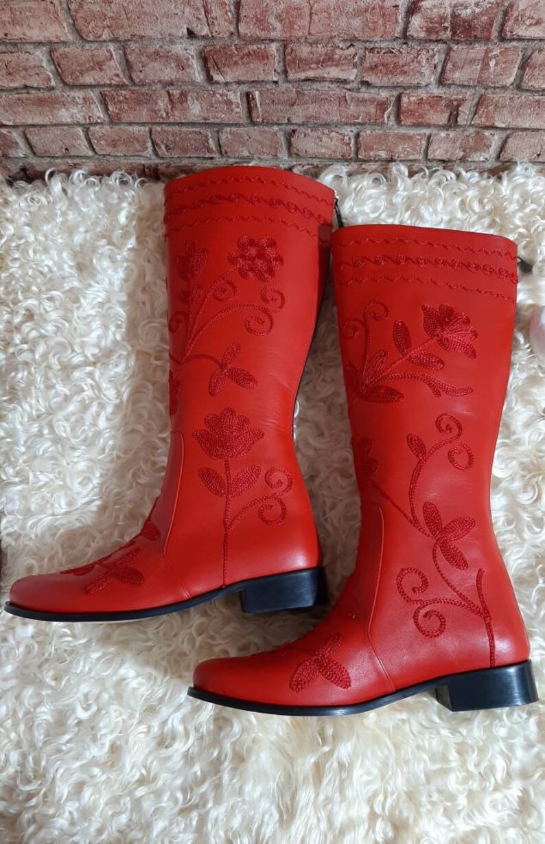 Puede incluir: Un par de botas rojas brillantes hasta la rodilla. Las botas presentan intrincados bordados florales en hilo rojo m&aacute;s oscuro. Tienen un tac&oacute;n bajo y negro y se muestran sobre una superficie blanca y esponjosa.