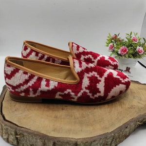 May include: A pair of red and white patterned velvet loafers with a brown leather sole. The loafers are on a wooden surface.