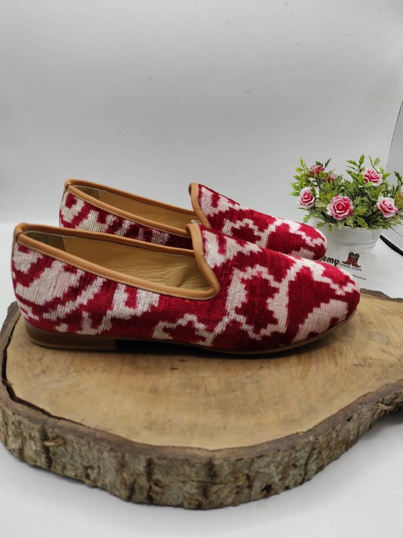 May include: A pair of red and white patterned velvet loafers with a brown leather sole. The loafers are on a wooden surface.