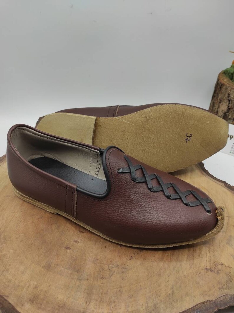 May include: A pair of brown leather slip-on shoes with a black braided design on the vamp. The shoes have a pointed toe and a flat sole. The size 37 is written on the sole of one shoe.