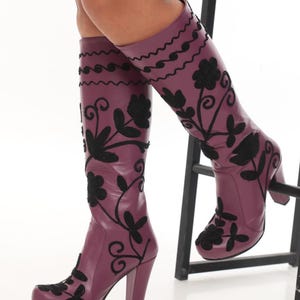 Puede incluir: Botas hasta la rodilla de color morado ciruela intenso con patrones florales y de enredaderas bordados en negro. Estas botas tienen una punta redondeada, un tac&oacute;n de bloque resistente y un acabado liso, llegando hasta la parte baja de la pantorrilla.