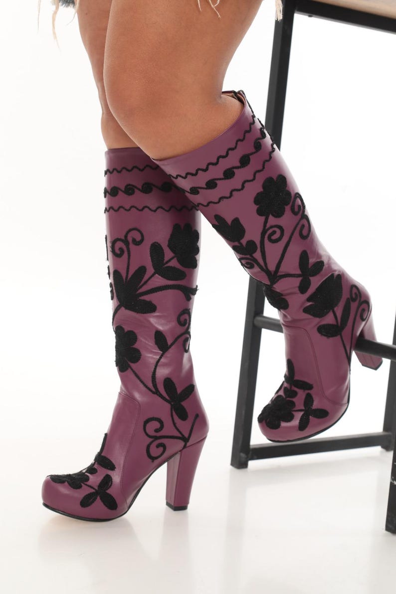 Puede incluir: Botas hasta la rodilla de color morado ciruela intenso con patrones florales y de enredaderas bordados en negro. Estas botas tienen una punta redondeada, un tac&oacute;n de bloque resistente y un acabado liso, llegando hasta la parte baja de la pantorrilla.