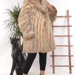 May include: A luxurious, knee-length fur coat in shades of tan and brown. The coat features a wide collar and long sleeves with fur cuffs. The model is wearing black leggings and black ankle boots. A decorative black panther statue is in the background.