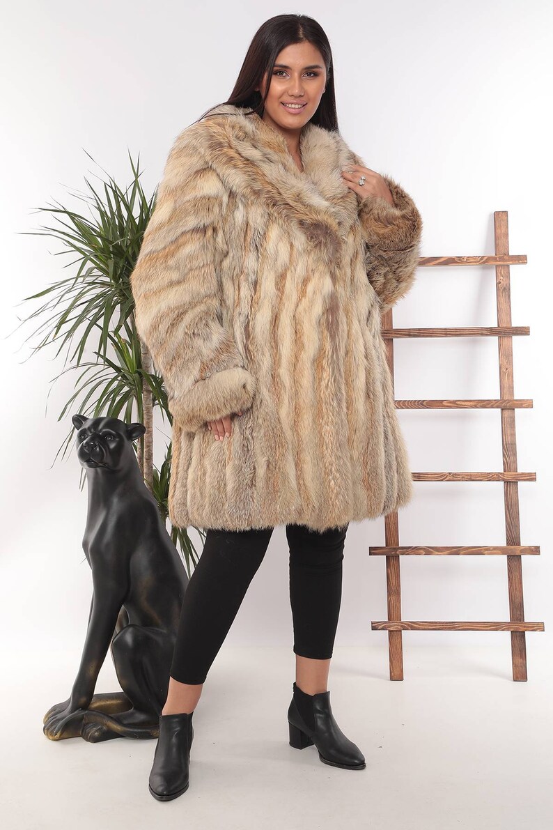 May include: A luxurious, knee-length fur coat in shades of tan and brown. The coat features a wide collar and long sleeves with fur cuffs. The model is wearing black leggings and black ankle boots. A decorative black panther statue is in the background.