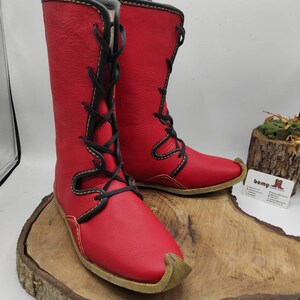 May include: A pair of red leather boots with black laces and a yellow stitch detail. The boots have a pointed toe and a flat sole.