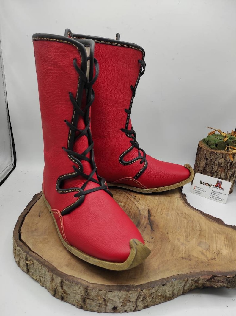 May include: A pair of red leather boots with black laces and a yellow stitch detail. The boots have a pointed toe and a flat sole.