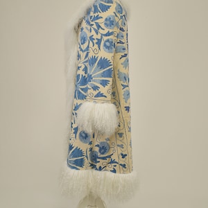 May include: A long white coat with blue floral embroidery. The coat has a cream fur collar and cuffs. The coat is displayed on a white mannequin against a white background.