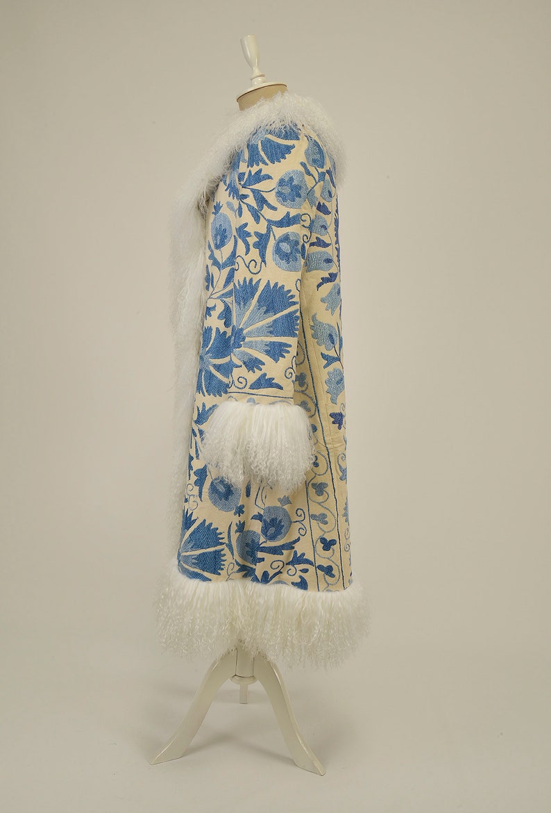 May include: A long white coat with blue floral embroidery. The coat has a cream fur collar and cuffs. The coat is displayed on a white mannequin against a white background.