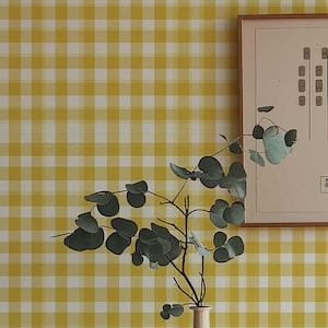 Può includere: Una cassettiera in legno in stile moderno di metà secolo con cassetti, un vaso bianco con vegetazione e opere d'arte incorniciate. Lo sfondo presenta una carta da parati a scacchi gialla e bianca. Una lampada a sospensione bianca e in legno pende dal soffitto.