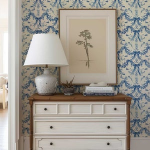K&ouml;nnte beinhalten: Eine wei&szlig;e Kommode mit drei Schubladen und braunen Holzbeinen. Die Kommode ist mit einer wei&szlig;en Lampe mit wei&szlig;em Lampenschirm, einem gerahmten Druck eines Baumes und einem Stapel B&uuml;chern dekoriert. Die Kommode steht vor einer Wand mit einer blau-wei&szlig;en Blumen-Tapete.