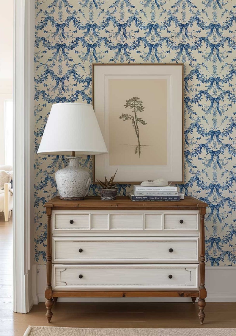 K&ouml;nnte beinhalten: Eine wei&szlig;e Kommode mit drei Schubladen und braunen Holzbeinen. Die Kommode ist mit einer wei&szlig;en Lampe mit wei&szlig;em Lampenschirm, einem gerahmten Druck eines Baumes und einem Stapel B&uuml;chern dekoriert. Die Kommode steht vor einer Wand mit einer blau-wei&szlig;en Blumen-Tapete.