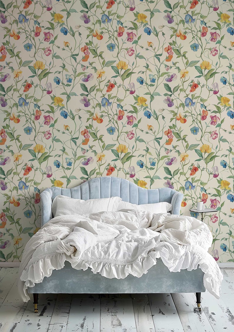 May include: A light blue upholstered bed with a scalloped headboard and white ruffled bedding, positioned against a cream-colored wallpaper featuring a repeating pattern of climbing vines with multi-colored sweet pea flowers in yellow, blue, and pink.