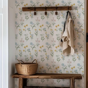 May include: A rustic wooden bench with a matching wall-mounted coat rack featuring four metal hooks. A woven wicker basket sits on the bench, and a light-colored jacket hangs from a hook. The wall is covered in white wallpaper with a floral pattern.
