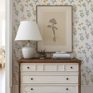 May include: A white wooden dresser with three drawers and dark knobs, topped with a white lamp, a small potted succulent, and a stack of books. Behind the dresser, a framed print of a tree hangs on a wall covered in floral wallpaper.