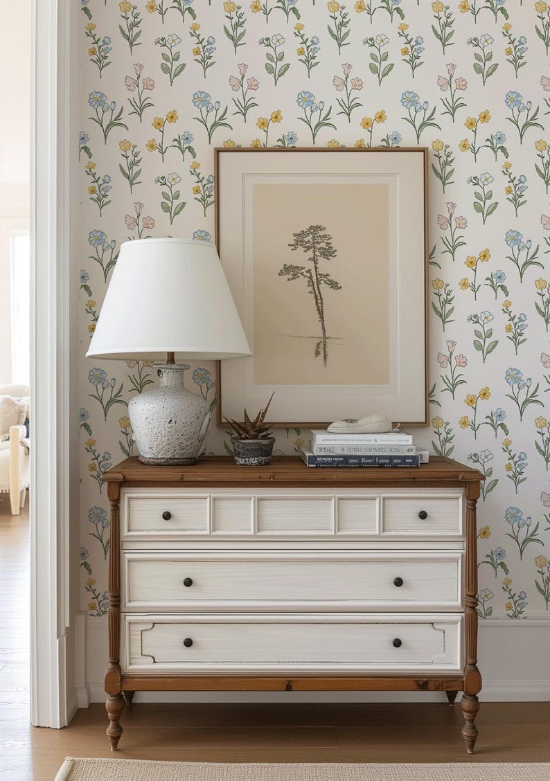 May include: A white wooden dresser with three drawers and dark knobs, topped with a white lamp, a small potted succulent, and a stack of books. Behind the dresser, a framed print of a tree hangs on a wall covered in floral wallpaper.