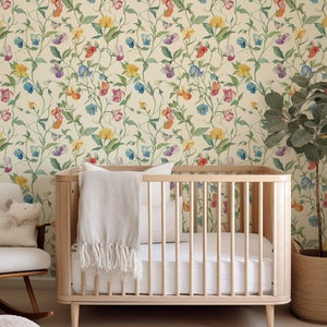 May include: A light-colored wooden crib with vertical slats, featuring a white mattress and a draped white throw blanket. The crib is positioned against a wall covered in wallpaper with a repeating pattern of colorful sweet pea flowers.