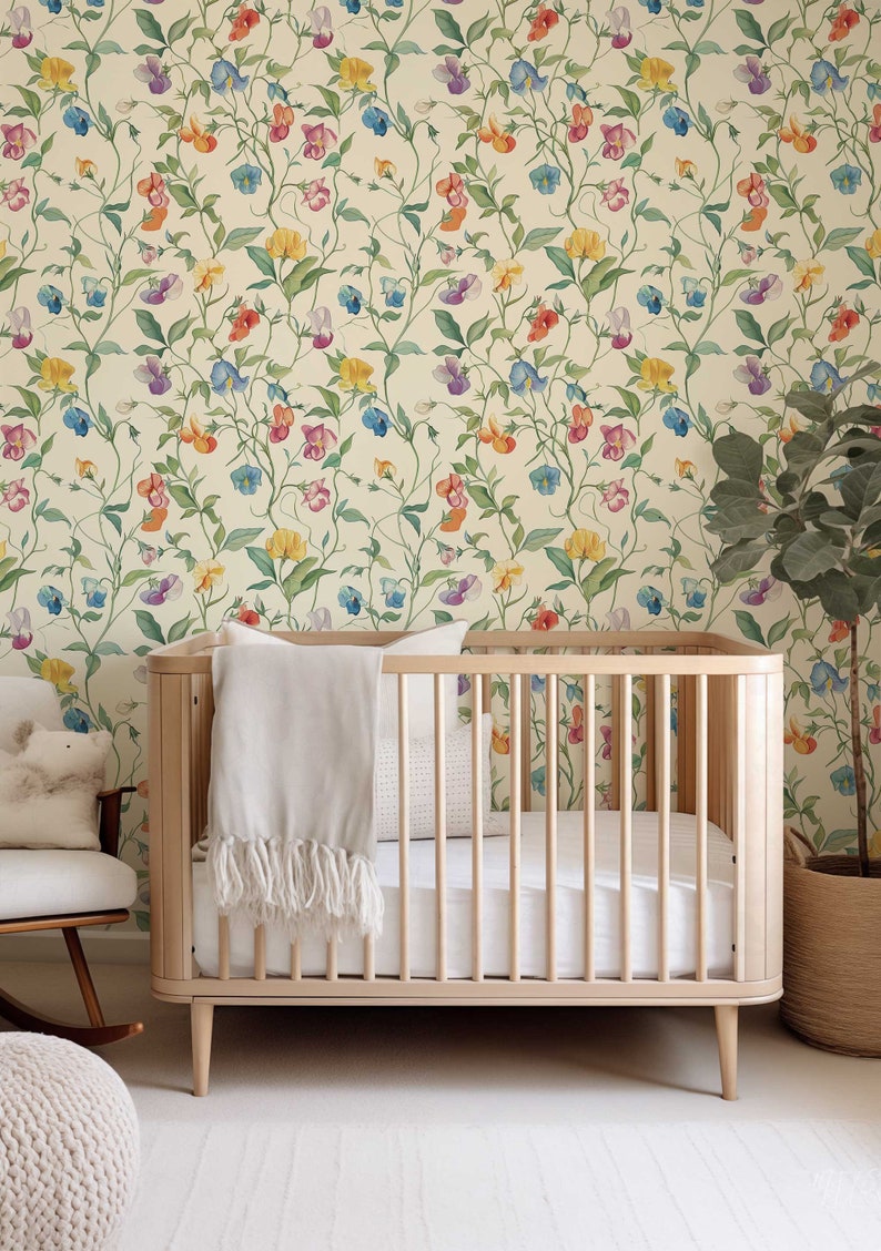 May include: A light-colored wooden crib with vertical slats, featuring a white mattress and a draped white throw blanket. The crib is positioned against a wall covered in wallpaper with a repeating pattern of colorful sweet pea flowers.