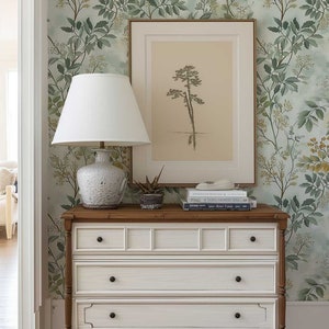 May include: A white dresser with three drawers and brown wooden legs. The dresser is decorated with a floral wallpaper featuring green leaves and yellow flowers. A white lamp with a white lampshade sits on top of the dresser, along with a framed print of a tree and a stack of books.