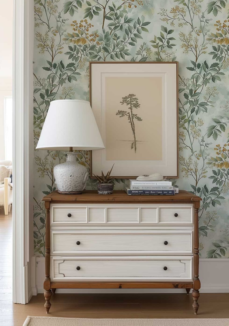 May include: A white dresser with three drawers and brown wooden legs. The dresser is decorated with a floral wallpaper featuring green leaves and yellow flowers. A white lamp with a white lampshade sits on top of the dresser, along with a framed print of a tree and a stack of books.