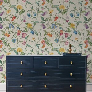 May include: A dark navy blue dresser with seven drawers, gold-toned hardware, and four gold-toned legs. The dresser is positioned against a wall covered in wallpaper featuring a repeating pattern of colorful sweet pea flowers and green vines.