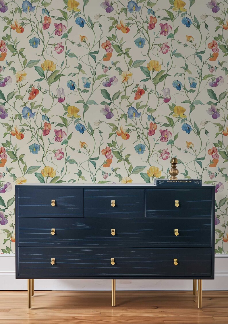 May include: A dark navy blue dresser with seven drawers, gold-toned hardware, and four gold-toned legs. The dresser is positioned against a wall covered in wallpaper featuring a repeating pattern of colorful sweet pea flowers and green vines.
