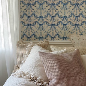 K&ouml;nnte beinhalten: Ein wei&szlig;es Bett mit einem wei&szlig;en Bettbezug und mehreren Kissen. Die Kissen sind in Wei&szlig;, Beige und Braun gehalten. Das Bett steht an einer Wand mit einer blauen und wei&szlig;en Blumen-Tapete.