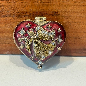 May include: A heart-shaped trinket box with a hinged lid. The box is red with a gold angel design and clear crystal accents. The box has a gold-colored metal frame and clasp.