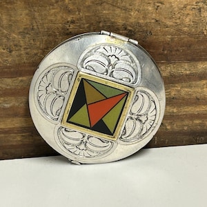 May include: A round, silver-toned compact with an ornate floral design. The center features a square with a geometric pattern in black, green, orange, and gold. The compact is open, revealing the interior.