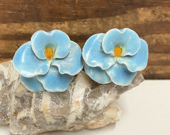 May include: A pair of blue ceramic flower earrings with yellow centers. The earrings are shaped like pansies and have a textured finish.