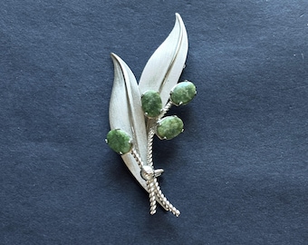 May include: A silver-toned metal brooch shaped like a plant stem with two elongated, textured leaves and three oval-shaped, mottled green stones set in prongs. The stems are twisted, and the leaves feature fine, etched line details.