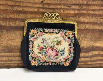 May include: A black coin purse featuring a central needlepoint panel with a colorful floral design of pink roses and small blue and yellow flowers. The purse has a gold-toned metal frame with an ornate, filigree-style clasp at the top.