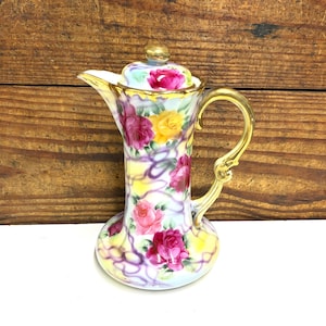 May include: A porcelain pitcher with a lid featuring a hand-painted floral pattern of pink, red, and yellow roses against a soft yellow and white background with purple accents. The pitcher has a gold-toned handle and a gold-trimmed rim.