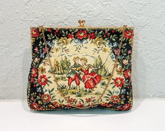 May include: A rectangular tapestry handbag featuring a central cream-colored panel with a scene of two figures dancing in a landscape, surrounded by a black border with red, yellow, and blue floral patterns, finished with a gold metal frame and chain.