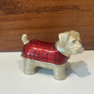 May include: A small, decorative dog figurine with a cream-colored body and a red, glittery coat. The dog is posed in a standing position, with its tail raised. The figurine is likely made of metal or ceramic.
