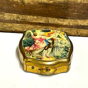 May include: A small, antique gold-toned metal box with a hinged lid. The lid features a colorful, detailed painting of a couple dancing, with a waterfall and cherubs in the background. The box is rectangular with rounded edges.