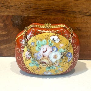 May include: A small, ornate purse with a gold chain strap. The purse is primarily red with gold accents and a yellow center panel. The center panel features a floral design with pink, white, and blue flowers.