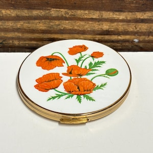 May include: A round, gold-toned metal compact mirror featuring a white enamel lid decorated with a vibrant illustration of orange poppy flowers and green leaves. The compact has a polished gold metal edge and a small latch for opening the case.