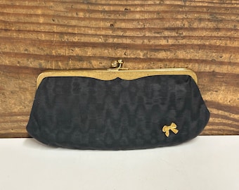 May include: A black clutch with a gold-colored clasp and a small bow accent. The clutch has a textured pattern and a gold-colored bow embellishment. The background is a wooden surface.