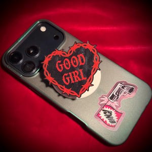 May include: A cell phone with a green case featuring two stickers. One sticker is a black heart with red barbed wire and the words "GOOD GIRL" in red. The other sticker is a gun with a "BANG!" flag.