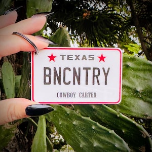 May include: A white Texas license plate keychain with a red border. The plate reads "TEXAS" above "BNCNTRY" and "COWBOY CARTER" in a bold font. Two red stars flank the word "TEXAS".
