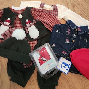 May include: A collection of children's clothing including a red plaid shirt with a snowman vest, a dark blue denim shirt with floral embroidery, a dark green beanie with a white pom-pom, a red beanie, and a boxed set of red and white shoes.