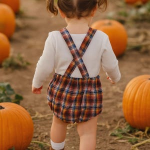 May include: A toddler wearing a white long-sleeved shirt and a plaid romper with suspenders. The romper features a pattern of orange, blue, and brown. The child is wearing brown boots and white socks, walking in a pumpkin patch.