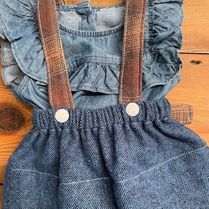 May include: A blue denim dress with a ruffled top and a plaid fabric strap. The dress has a matching blue and black herringbone skirt with white buttons.