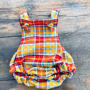 May include: A colorful plaid baby romper in shades of red, yellow, orange, and blue. The garment features shoulder straps with decorative knots and a gathered bottom. The fabric appears to be a soft flannel.