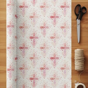 May include: A roll of wrapping paper with a repeating pattern of watercolor crosses in shades of pink and red, adorned with floral and heart accents. A pair of black scissors, twine, and tape are on a wooden surface.