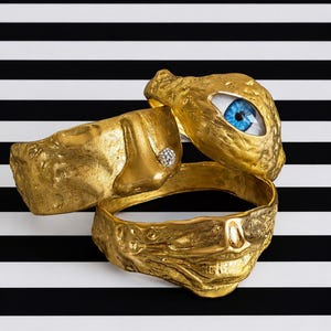 Può includere: Due bracciali rigidi in metallo dorato con superfici testurizzate e organiche. Uno presenta un inserto a forma di occhio blu realistico e l'altro un naso scolpito con un piccolo cristallo scintillante. Le superfici hanno una finitura grezza.